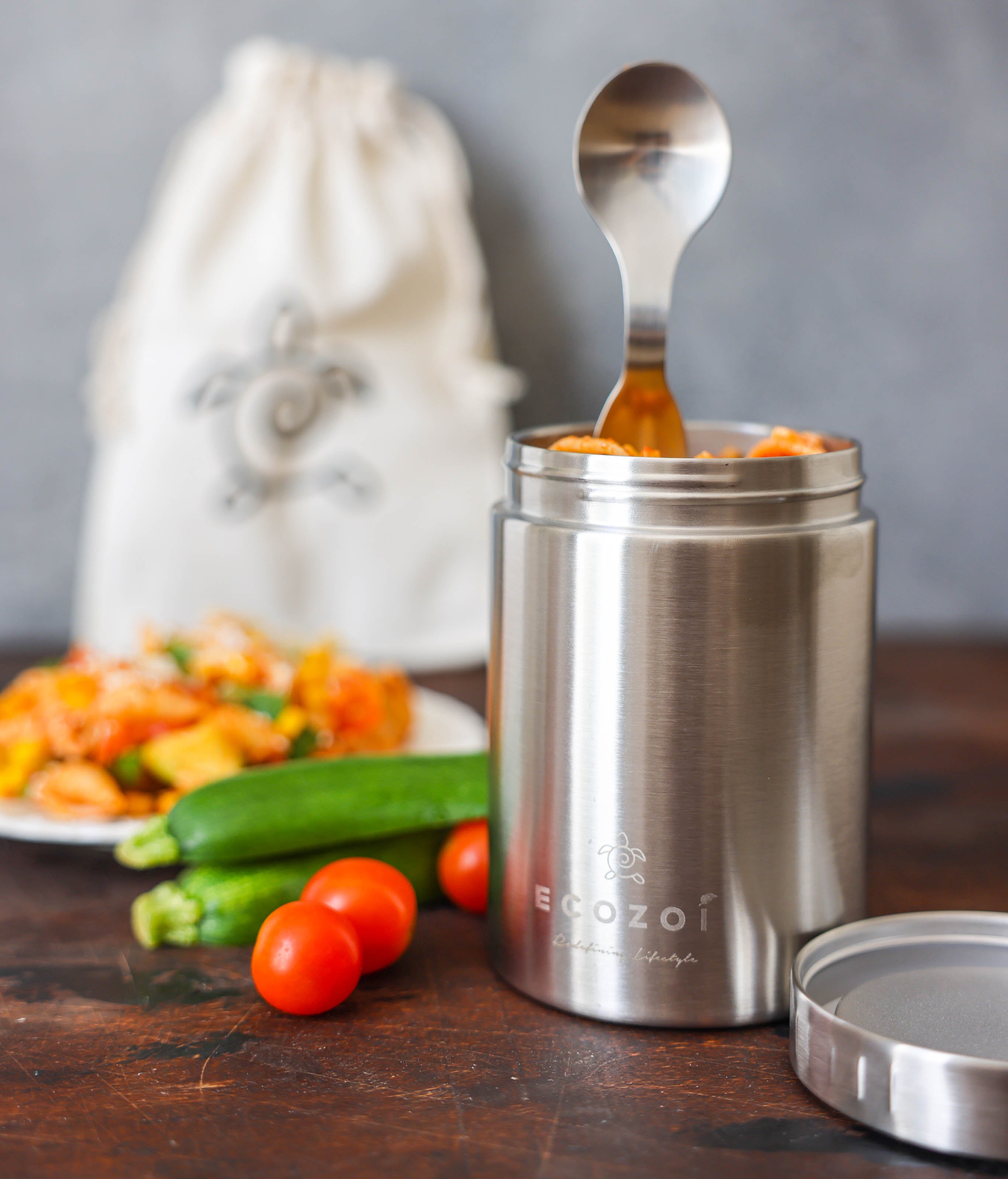Stainless steel food container with spoon, vegetables on a table, and blurred background