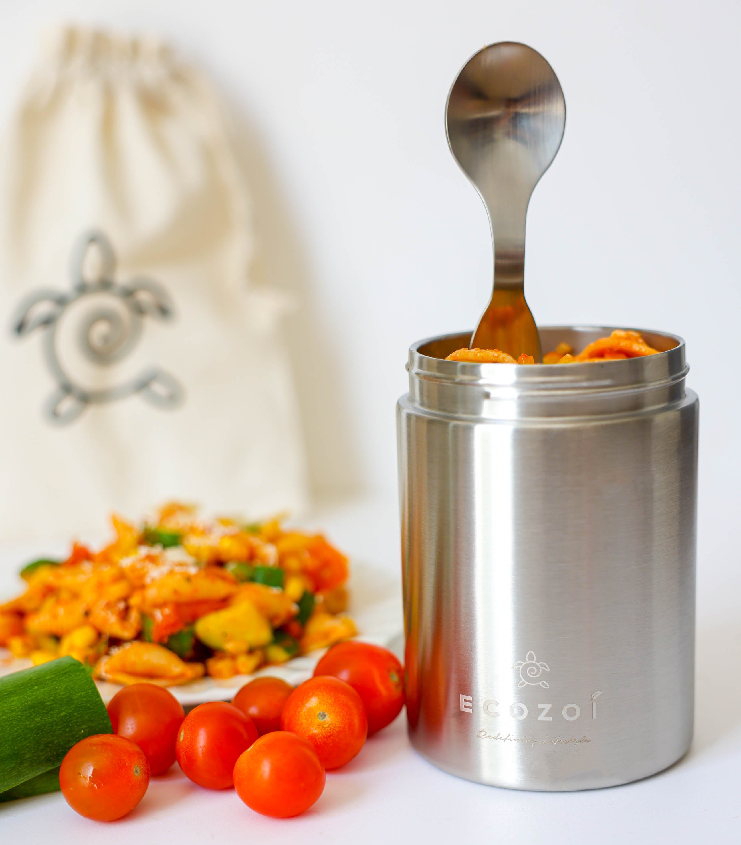 Stainless steel container with a spoon, surrounded by tomatoes and salad, on a white background.