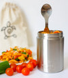 Stainless steel container with a spoon, surrounded by tomatoes and salad, on a white background.