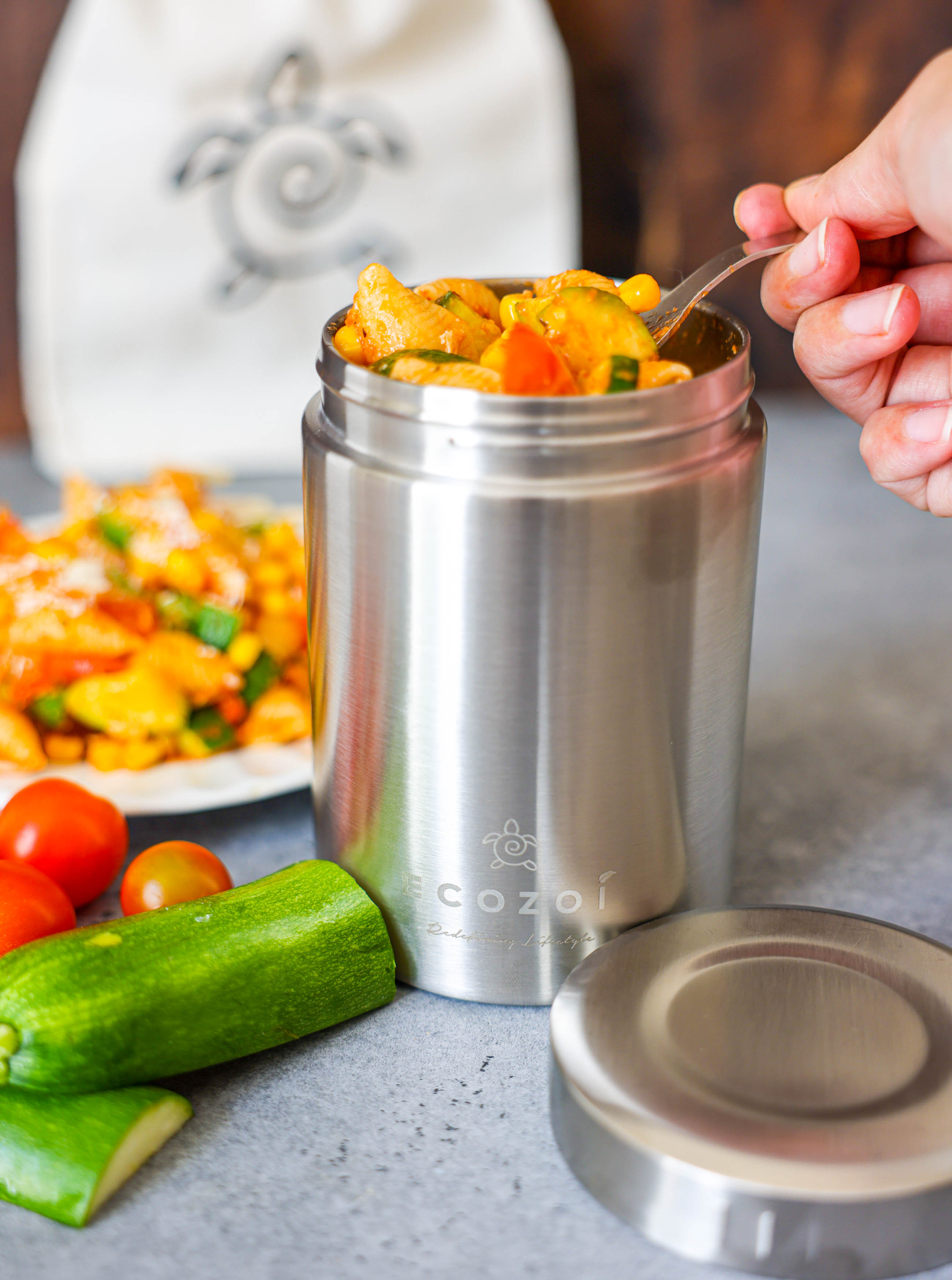 Stainless steel food container with a spoonful of food, surrounded by vegetables and a blurred dish in the background.