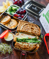 Lunchbox with a sandwich, cookies, fruits, and a container of hummus on a wooden surface.