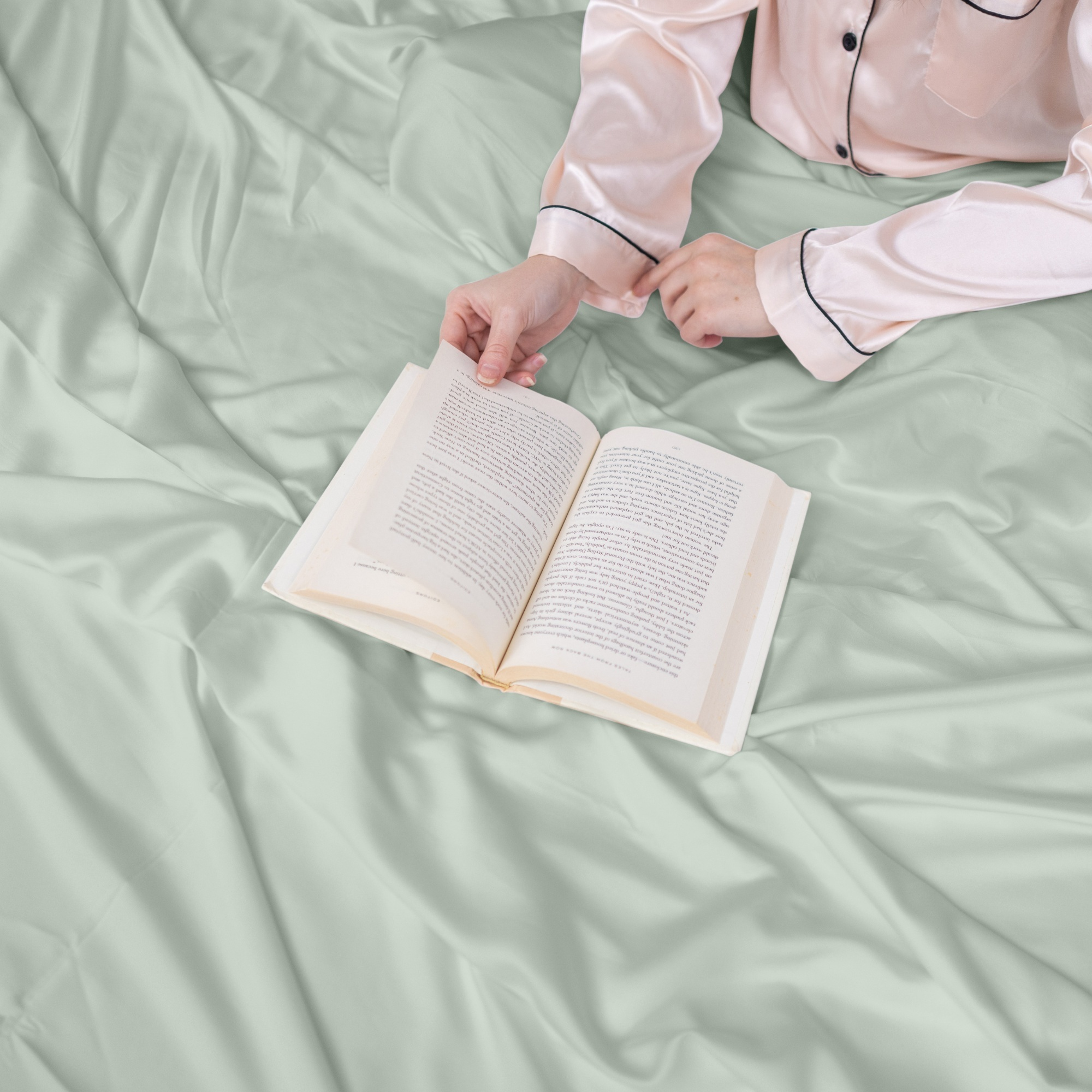 Person in pajamas holding an open book on a light green bedspread