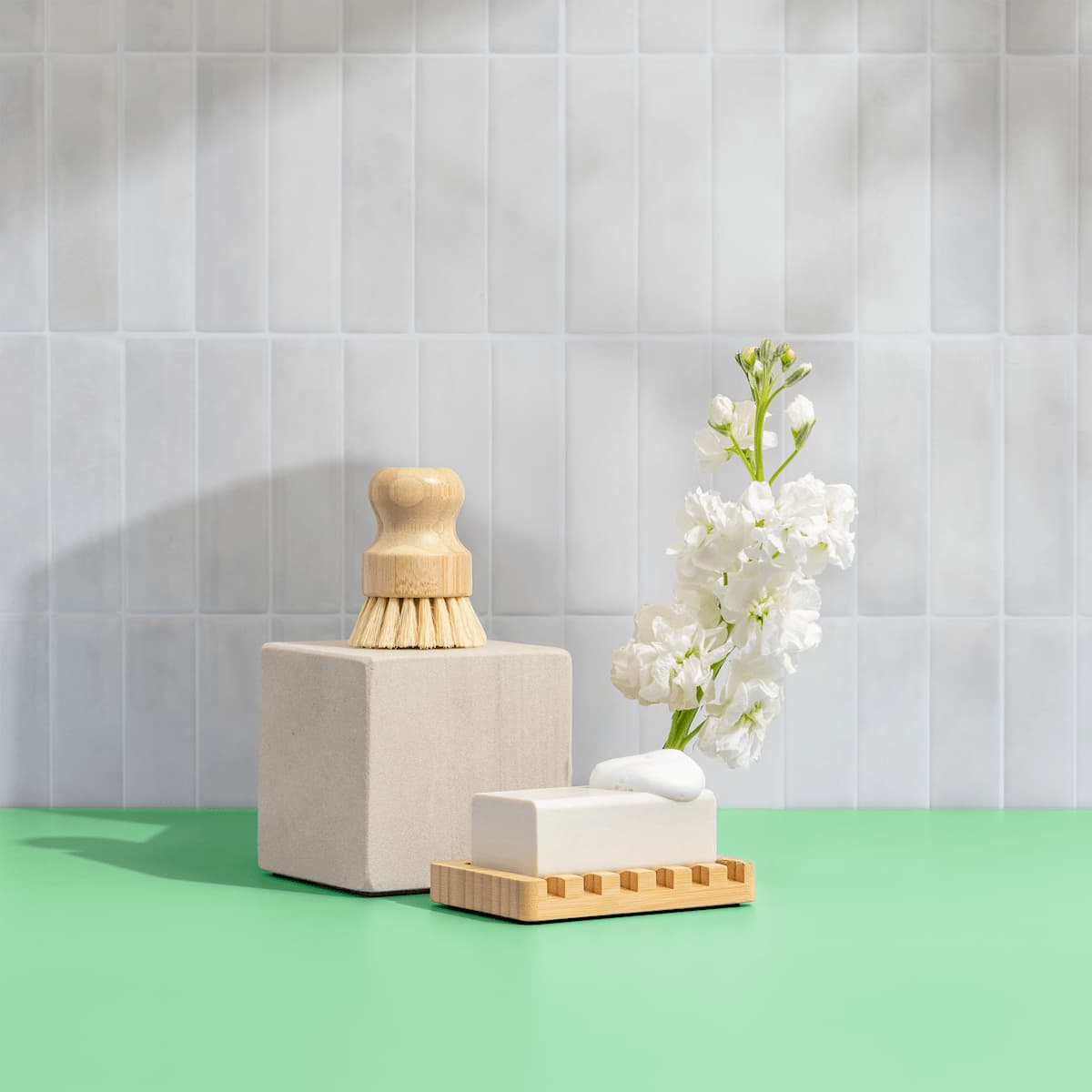 Bathroom setting with a scrub brush, soap, and flowers on a green surface against a tiled wall.