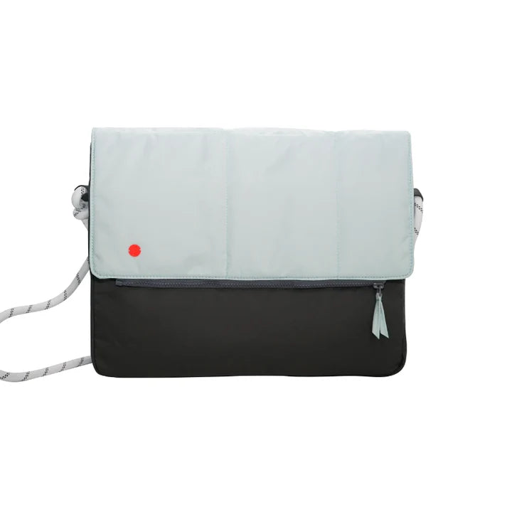 A two-tone messenger bag with a light mint green top flap and a black bottom body, featuring a small red dot logo on the flap, a gray and white patterned shoulder strap, and a tassel zipper pull on a white background.