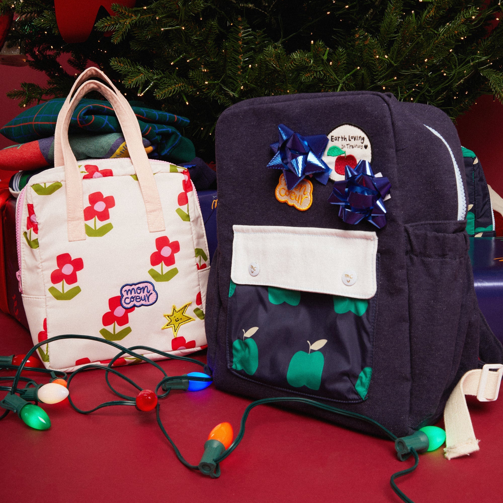 Floral-patterned handbag and navy backpack with apple design on a red surface with Christmas lights.