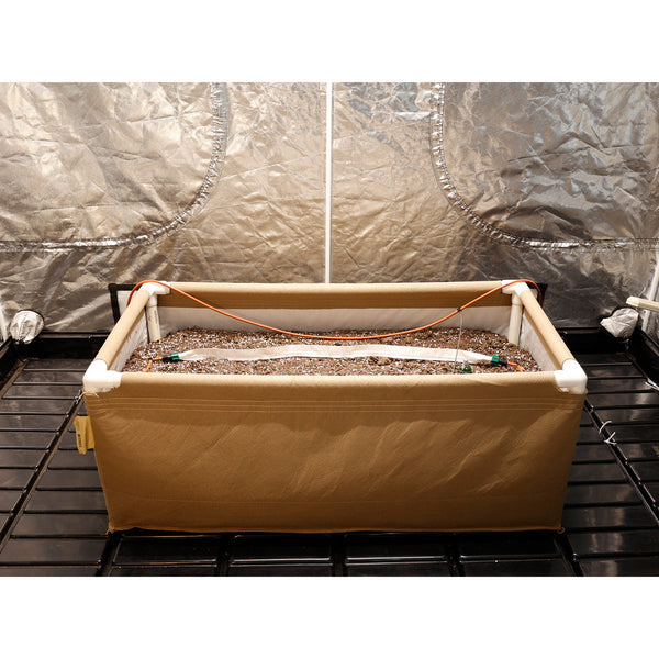 Rectangular grow box with soil and irrigation system inside, against a reflective wall.