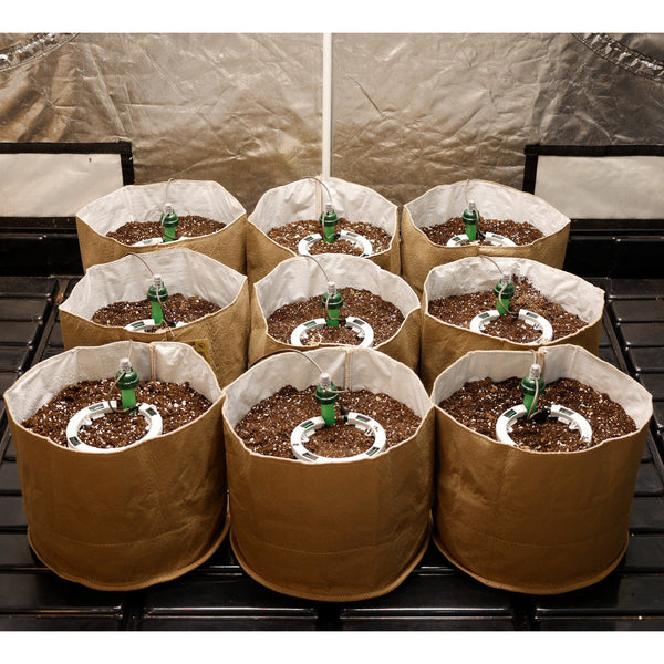 Brown grow bags with soil and green bottles on a metal rack against a beige tarp.