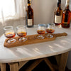 Wooden whiskey tasting tray with four glasses of whiskey on a table with bottles in the background.