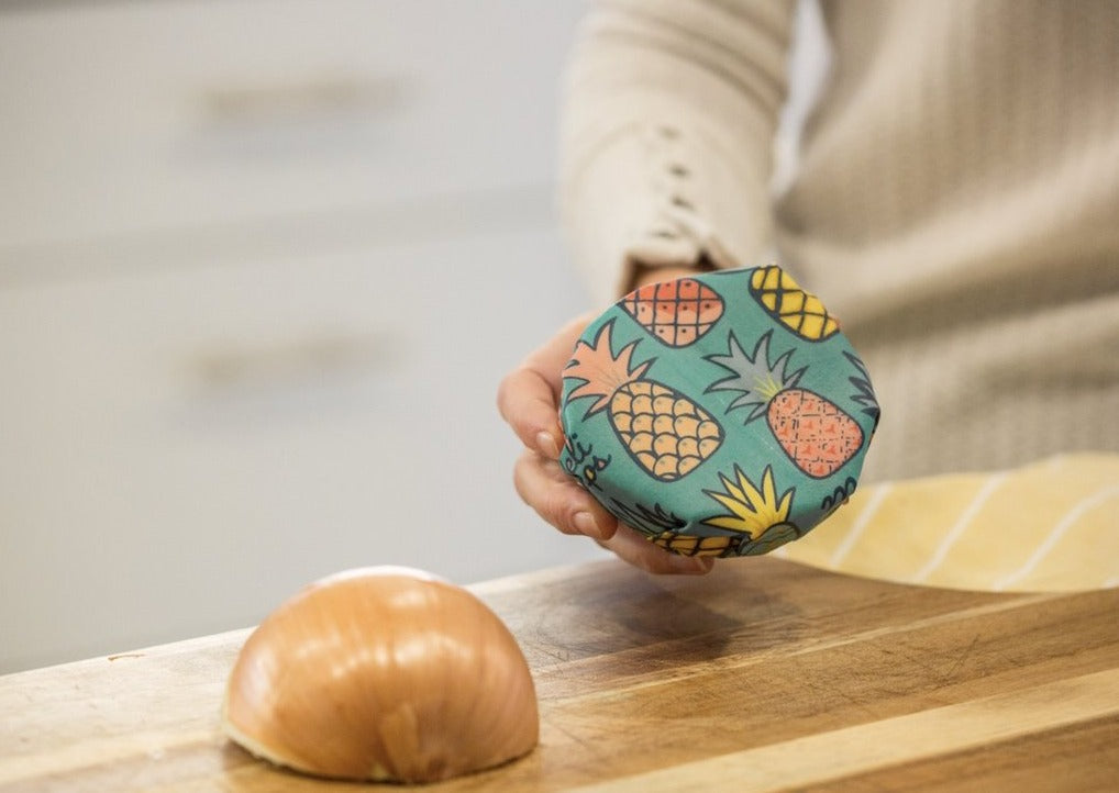 Person holding a pineapple-patterned food storage container on a wooden cutting board with an onion.