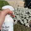 Person holding a bottle of 'Elm Dirt Shield' in front of a bush