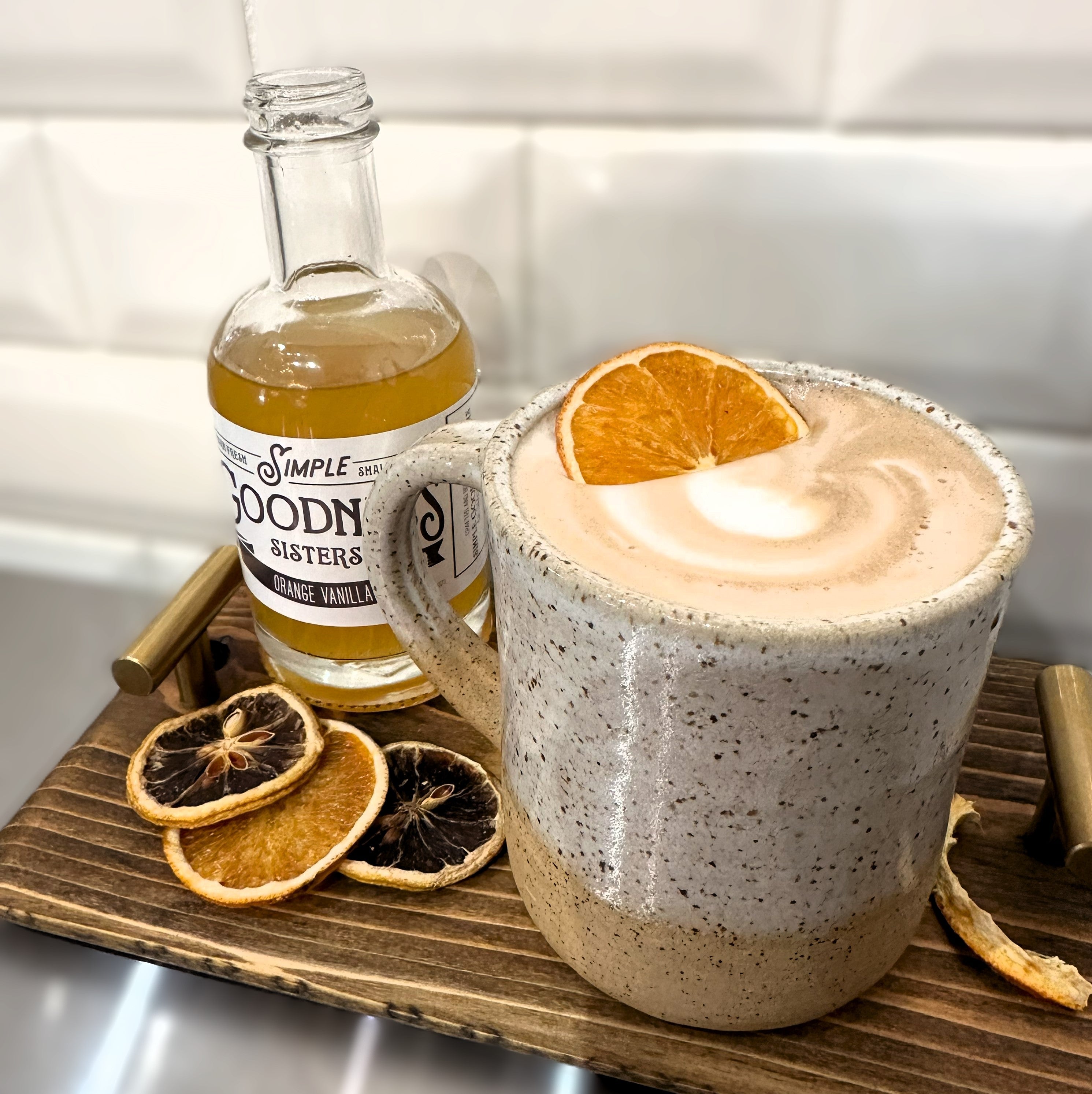 A speckled ceramic mug filled with a frothy latte topped with an orange slice, set on a wooden tray beside a glass bottle labeled Simple Woodn Bisters and dried orange slices.