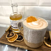 A speckled ceramic mug filled with a frothy latte topped with an orange slice, set on a wooden tray beside a glass bottle labeled Simple Woodn Bisters and dried orange slices.