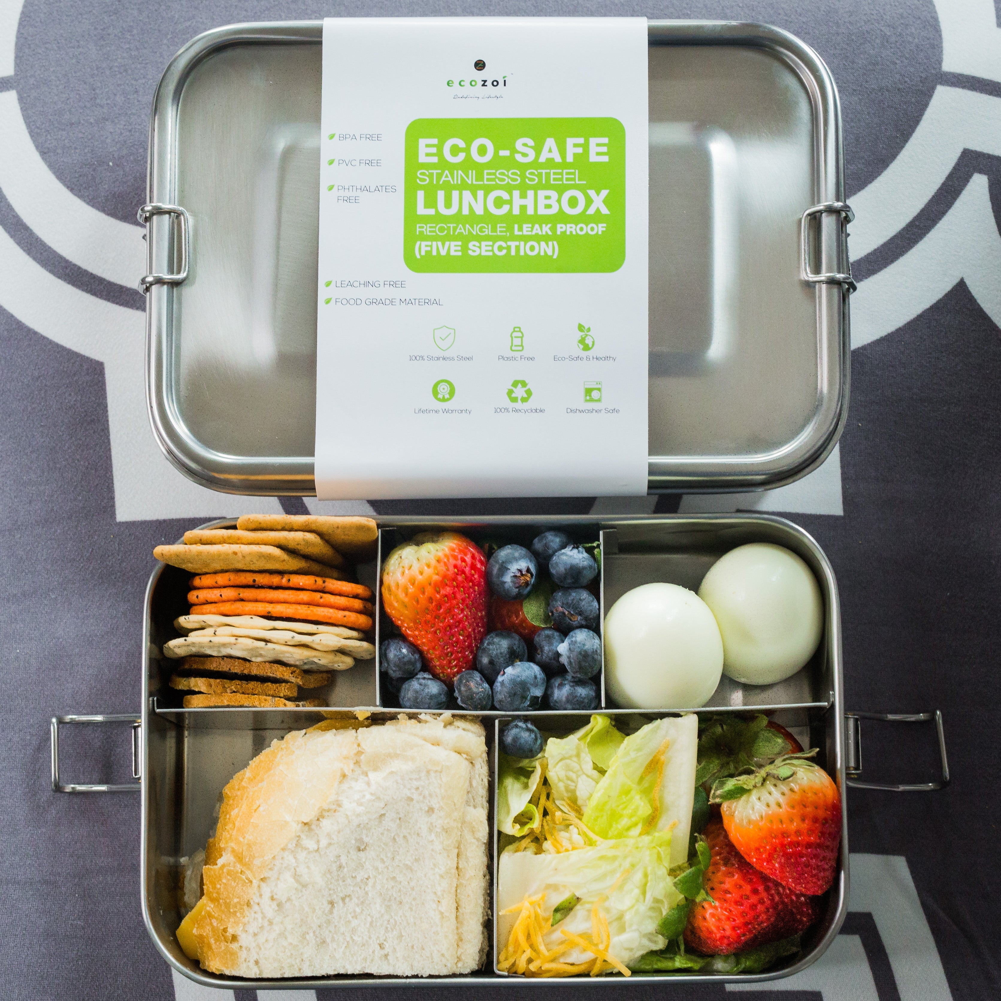 Metal lunchbox with compartments filled with food and an eco-safe label on a patterned background