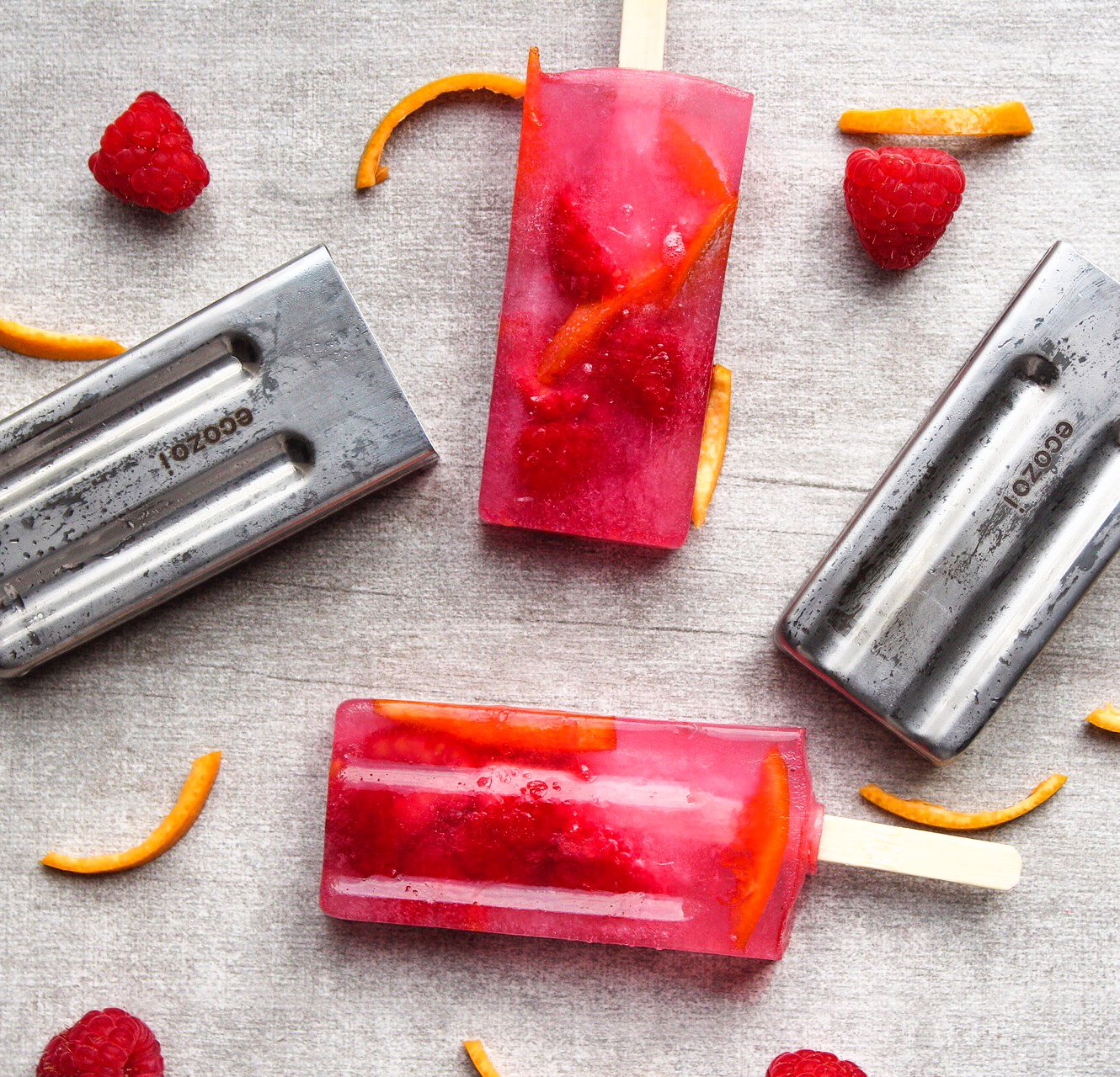 Two red popsicles with orange slices and raspberries on a gray surface, surrounded by popsicle molds.