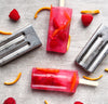 Two red popsicles with orange slices and raspberries on a gray surface, surrounded by popsicle molds.