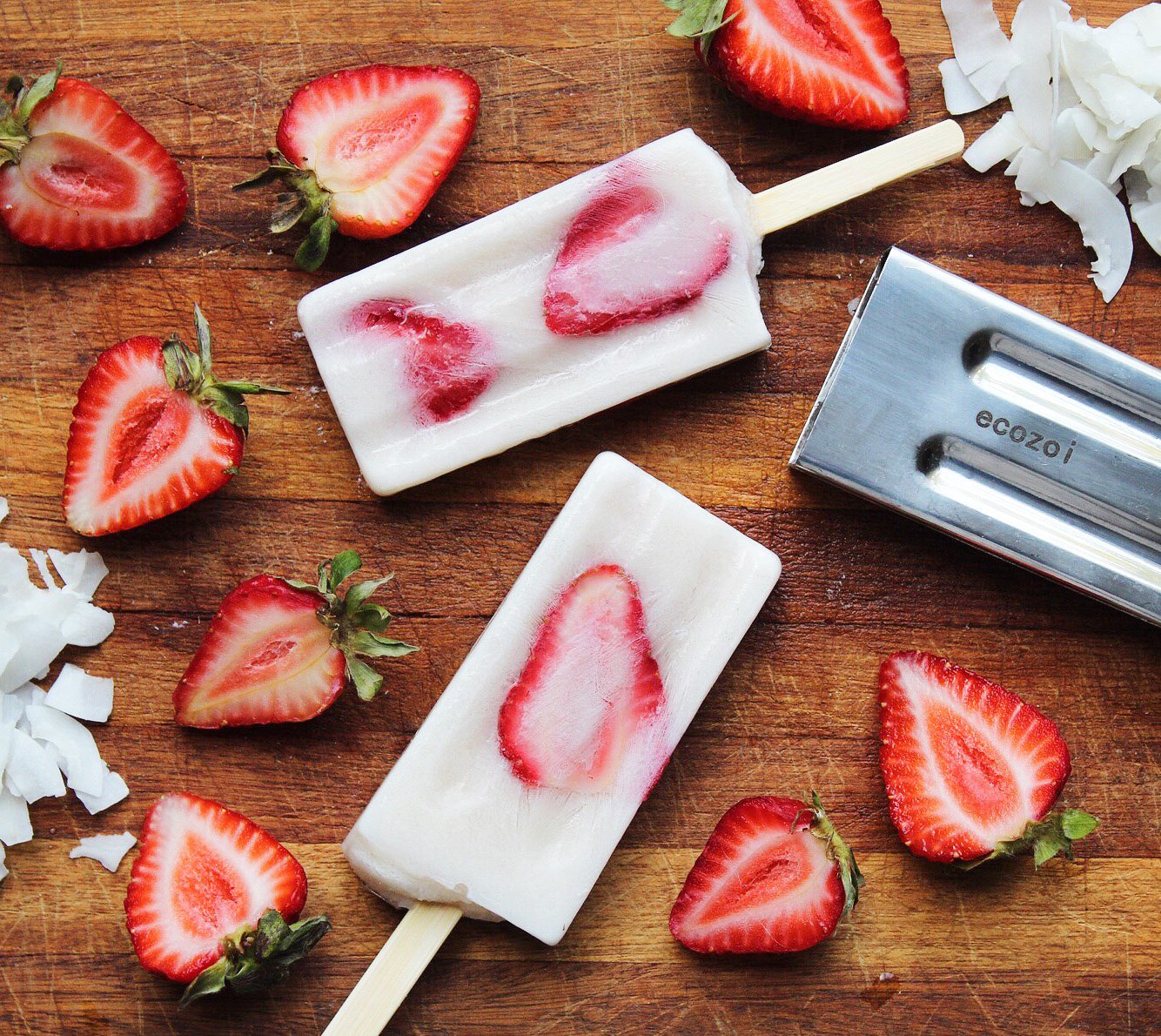 Two strawberry popsicles on a wooden surface with sliced strawberries and coconut flakes.
