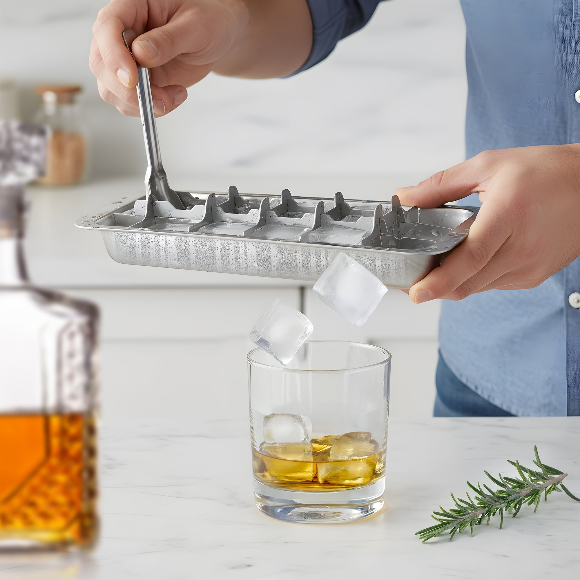 Person using a metal ice cube tray to fill a glass with ice cubes on a kitchen counter.