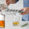 Person using a metal ice cube tray to fill a glass with ice cubes on a kitchen counter.