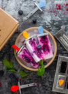 Two pink popsicles with blueberries on a wooden plate, surrounded by berries and a cardboard box.