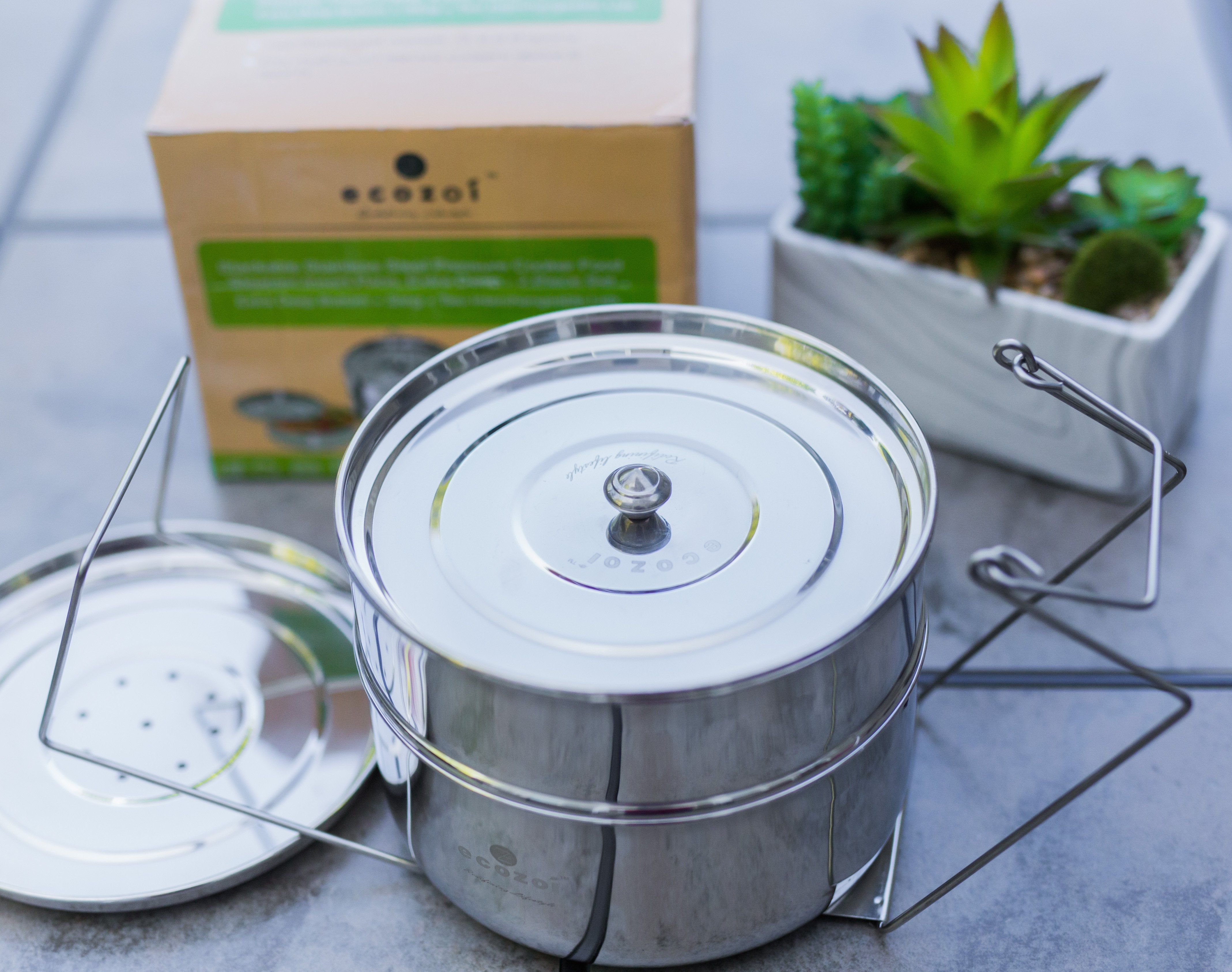 Stainless steel container with lid on a gray surface, next to an ecozai box and plant.