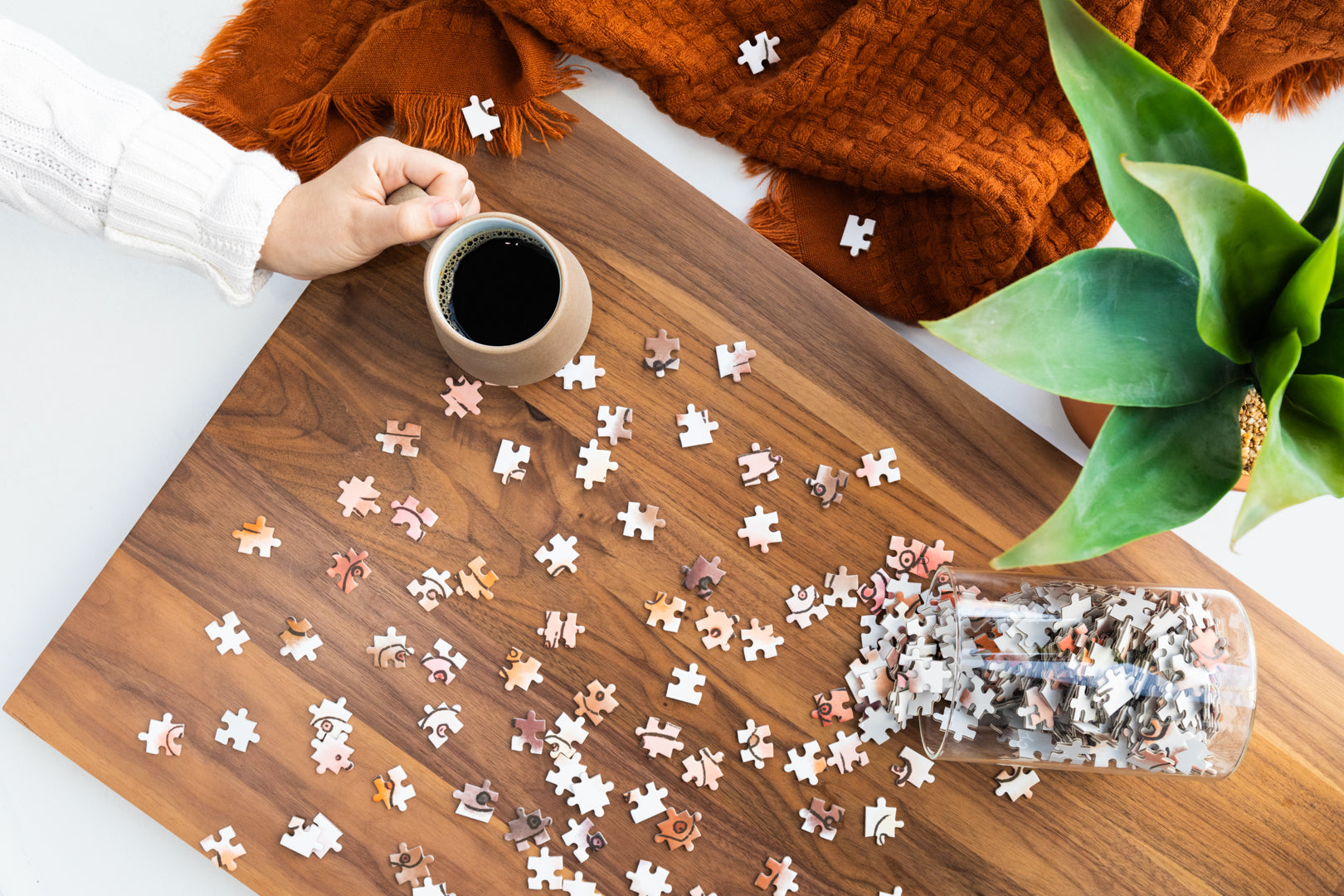 Puzzle pieces scattered on a wooden table with a hand holding a mug of coffee and a plant in the background.