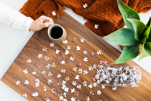 Puzzle pieces scattered on a wooden table with a hand holding a mug of coffee and a plant in the background.
