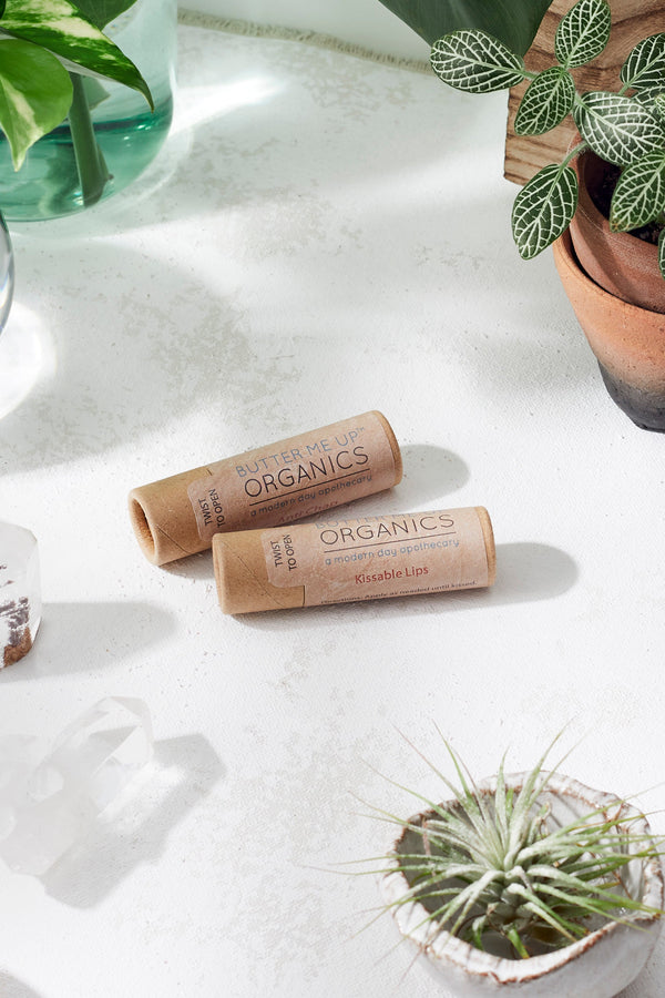 Two organic lip balm tubes on a white surface with plants in the background