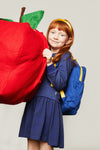 A young girl with red hair wearing a navy blue dress, yellow headband, and blue backpack with yellow straps, holding a giant red apple plush toy against a light gray studio background.