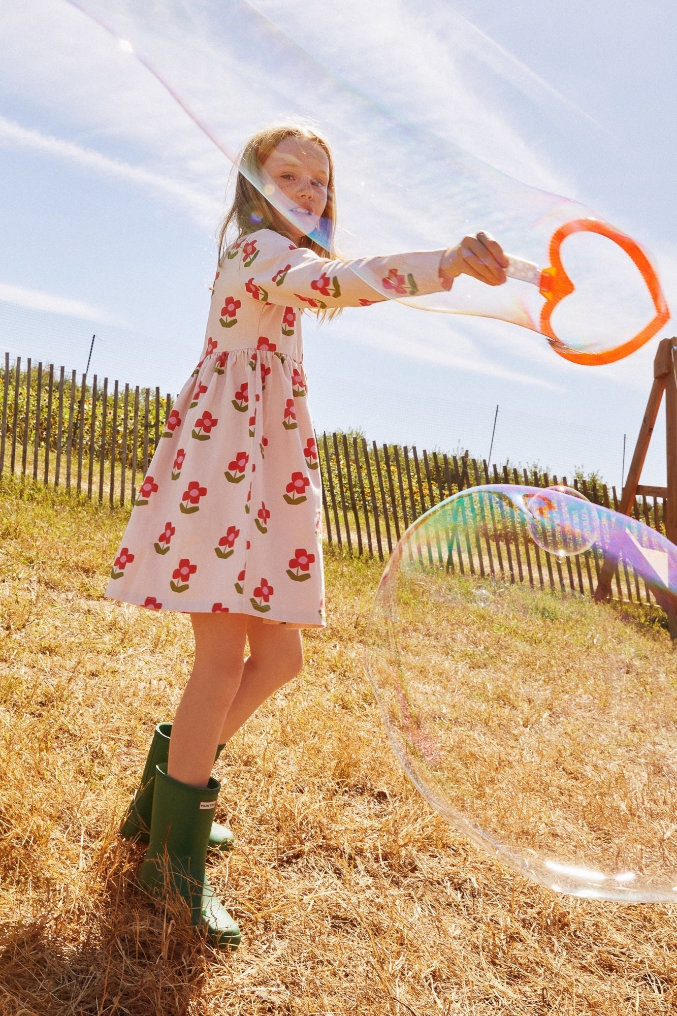 Child in a floral dress and green boots blowing bubbles outdoors on a sunny day.