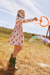 Child in a floral dress and green boots blowing bubbles outdoors on a sunny day.