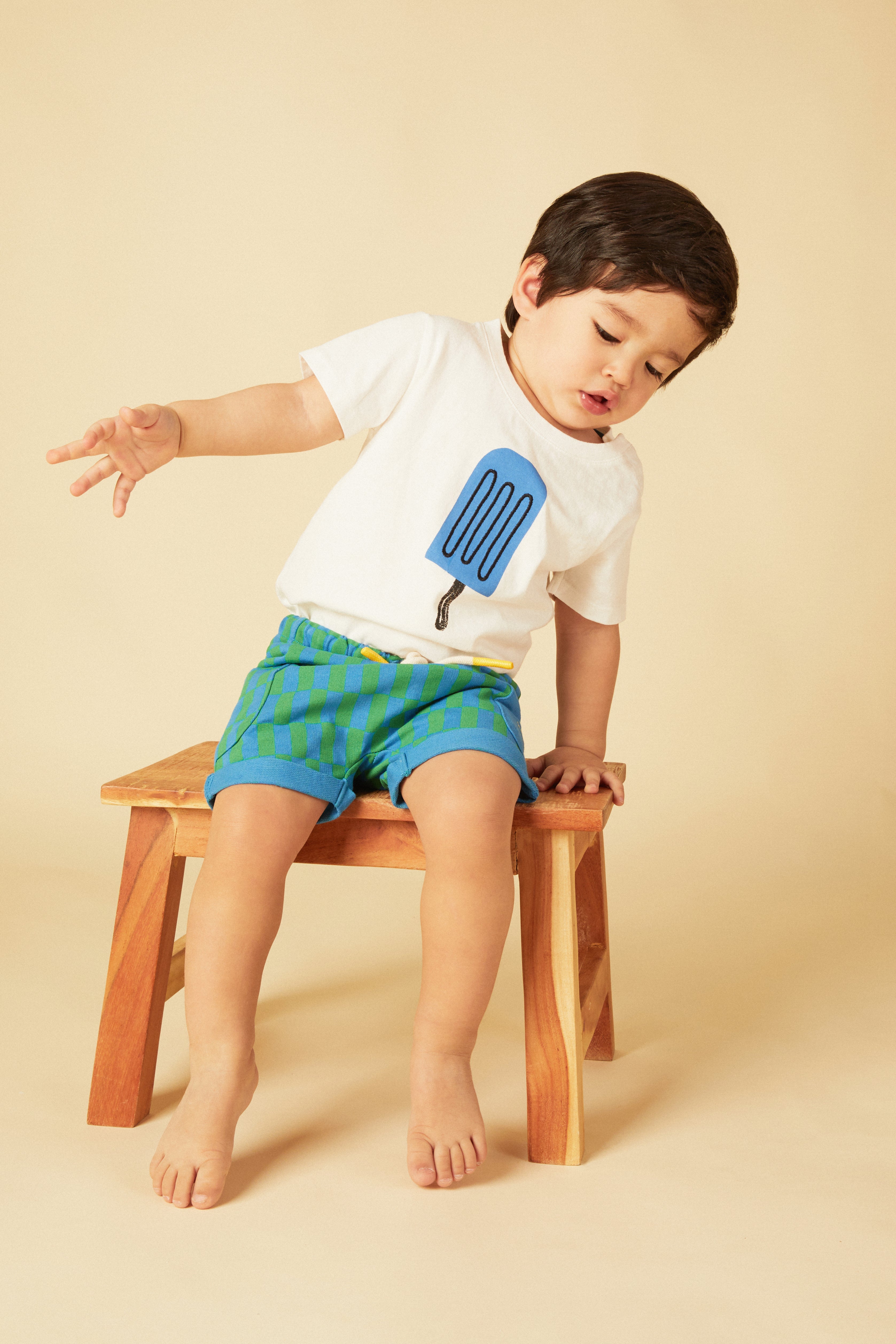 Child sitting on a wooden stool wearing a white shirt with a blue ice cream cone design and blue shorts.