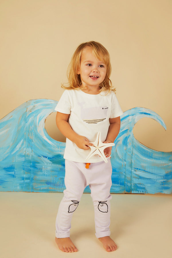 Child wearing a white t-shirt and light-colored pants with a starfish design, holding a starfish in front of a blue wave painting.