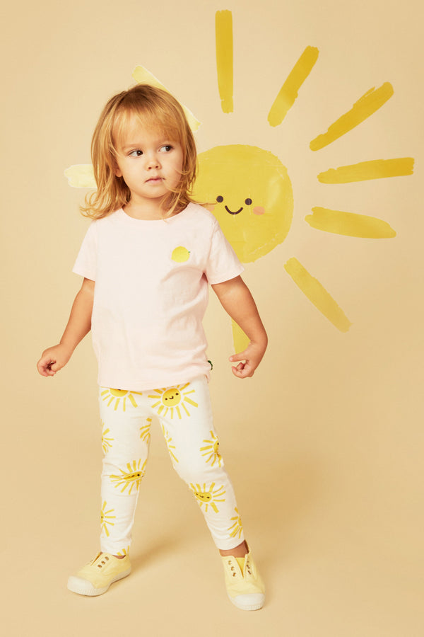 Child wearing a light pink shirt and white pants with yellow sun patterns, standing against a beige background with a yellow sun illustration.