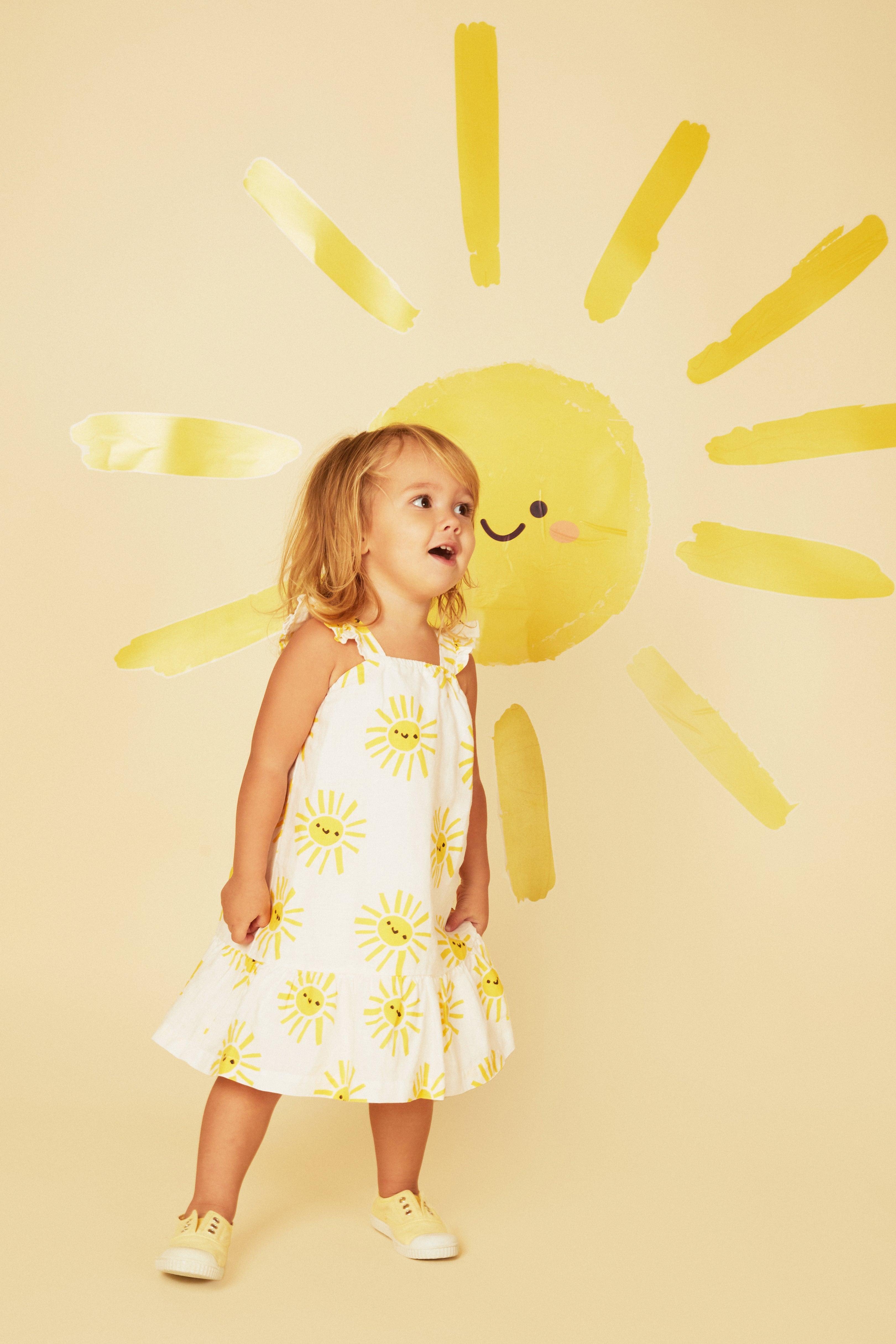 Child wearing a yellow and white dress with floral patterns against a yellow background with a sun design.