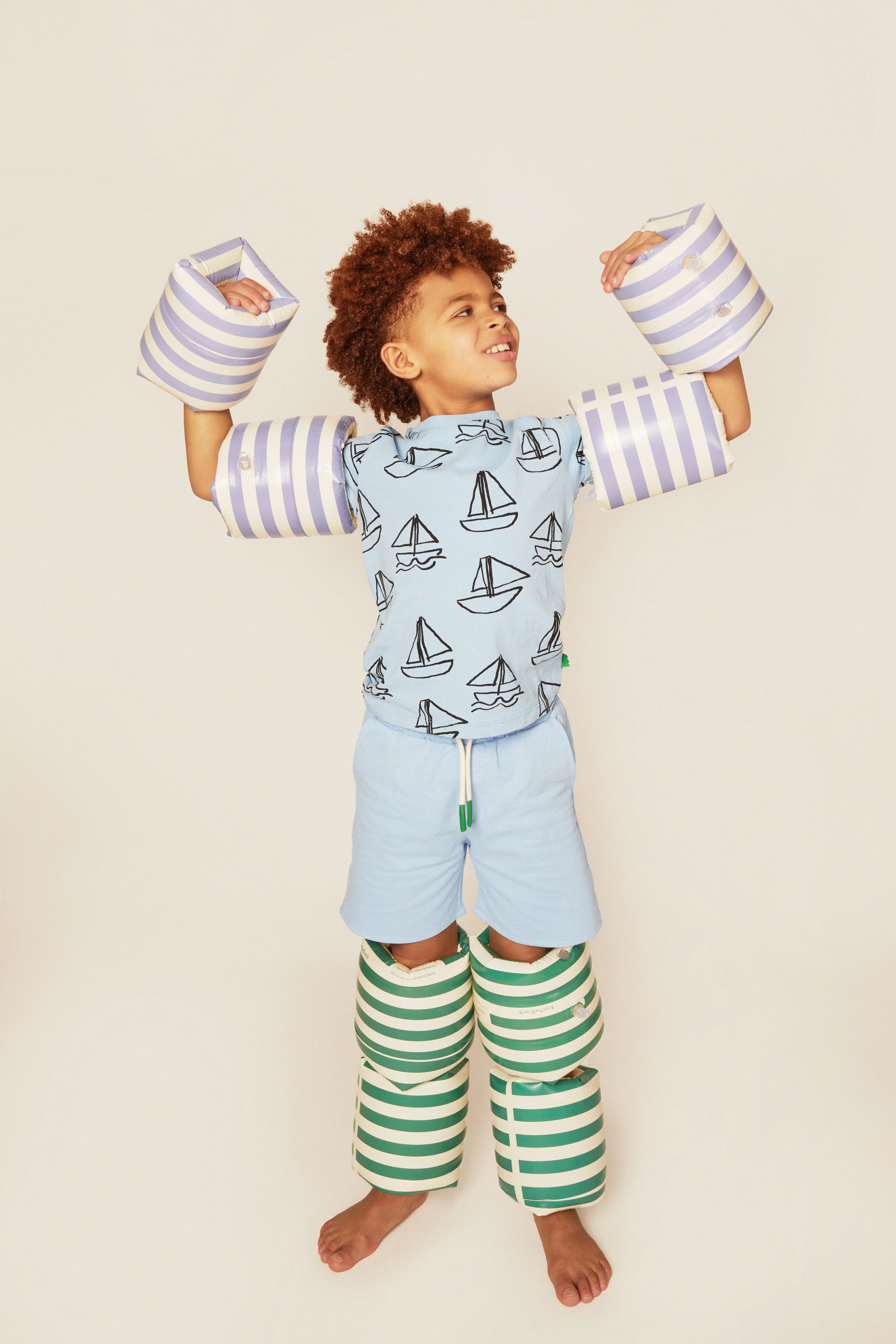 Child wearing a blue shirt with sailboat pattern, light blue shorts, green and white striped socks, and white shoes with blue stripes on a beige background.