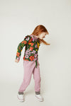 A young girl with red hair is shown from the side in a studio setting, wearing a dark green long-sleeve top with a colorful floral print, dusty pink sweatpants, and white sneakers.