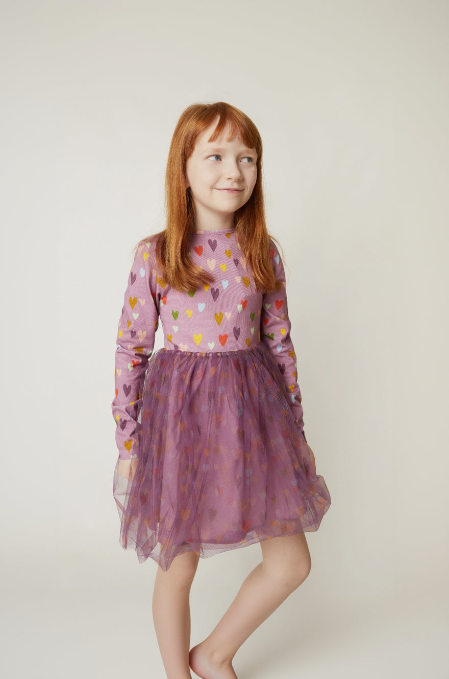 A young girl with red hair wearing a purple long-sleeve dress with a patterned bodice and a tulle skirt, standing against a plain light background.