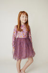 A young girl with red hair wearing a purple long-sleeve dress with a patterned bodice and a tulle skirt, standing against a plain light background.