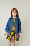 A young girl with red hair stands against a plain light beige studio background, wearing a blue denim jacket over a colorful floral dress with yellow, pink, green, and orange flowers.