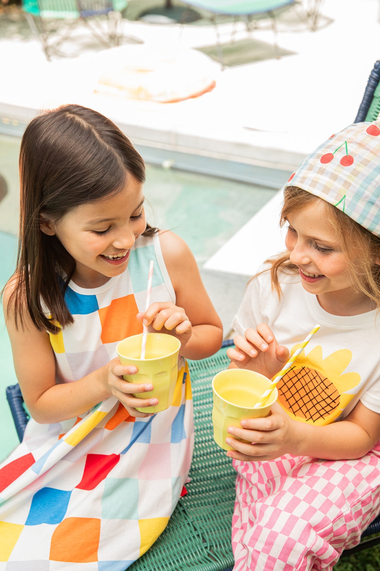 Two young girls sitting outdoors, holding yellow cups with straws.