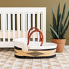 Wicker baby basket with white cushion in front of a white crib with a plant in the background