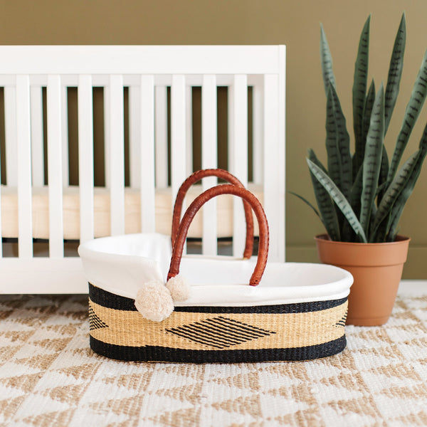 Wicker baby basket with white cushion in front of a white crib with a plant in the background