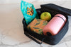 Black lunch bag with a green snack bag, pink thermos, and green apple on a marble surface