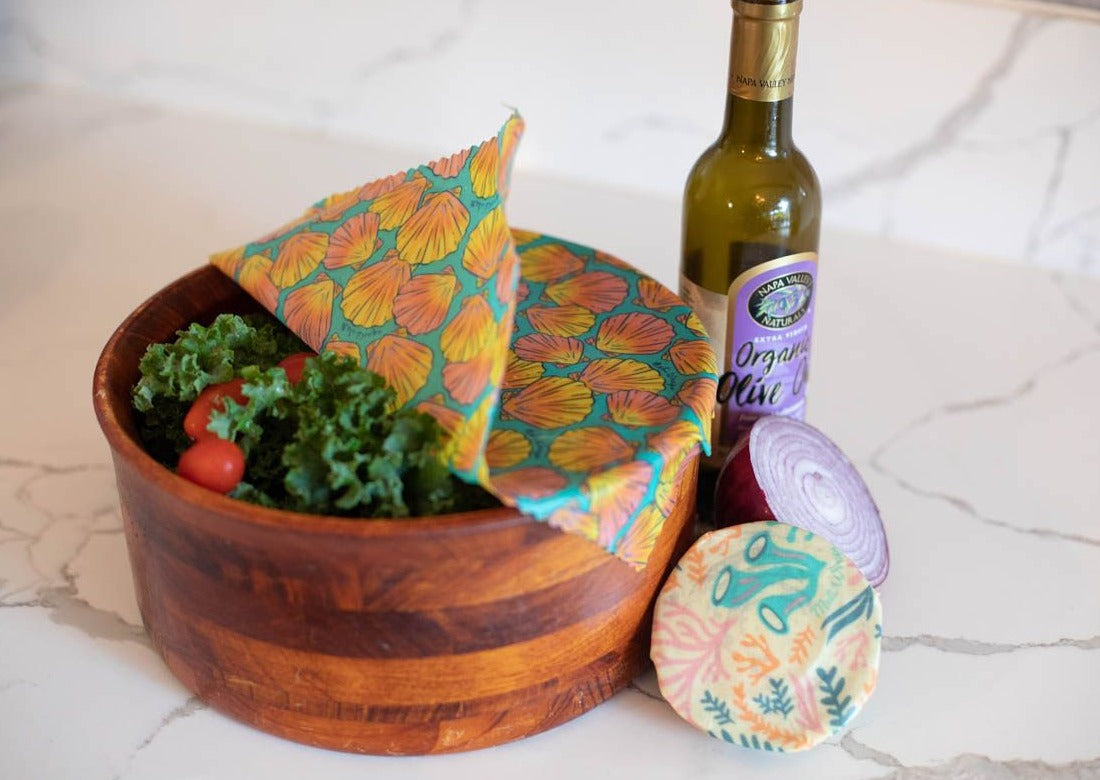 Wooden bowl with produce, a bottle of olive oil, and reusable food wraps on a marble surface.