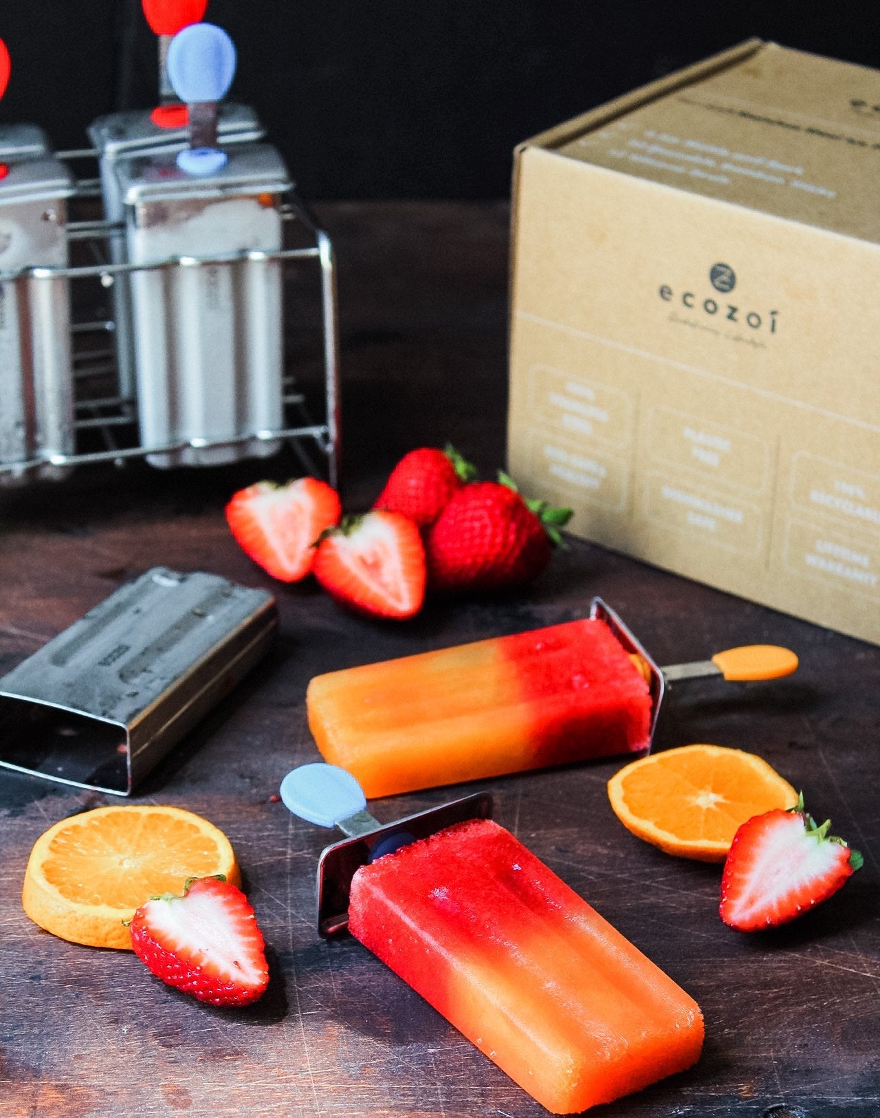 Two orange and red popsicles on a wooden surface with strawberries, oranges, and an ecozot box in the background.