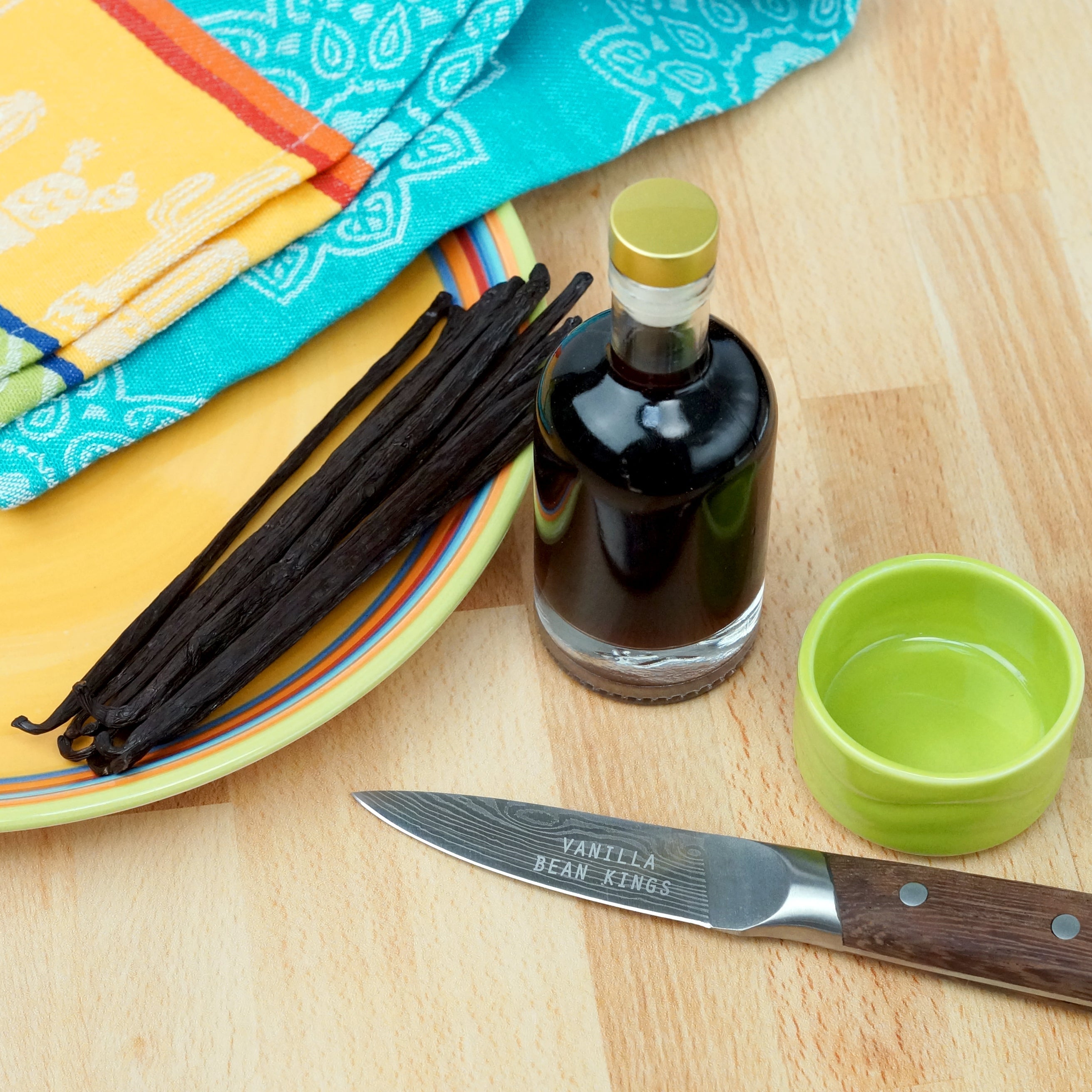 Vanilla beans, bottle of vanilla extract, and knife on a wooden surface with colorful cloths in the background.
