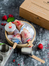 Ice cream popsicles with raspberry pieces in a wooden bowl on a dark surface, with an eco-friendly packaging box in the background.