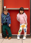 Two children standing against a red wall, one wearing a blue jacket and green pants, the other in a pink jacket and colorful pants.