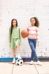 Two young girls standing next to each other with a basketball and soccer ball against a white brick wall.