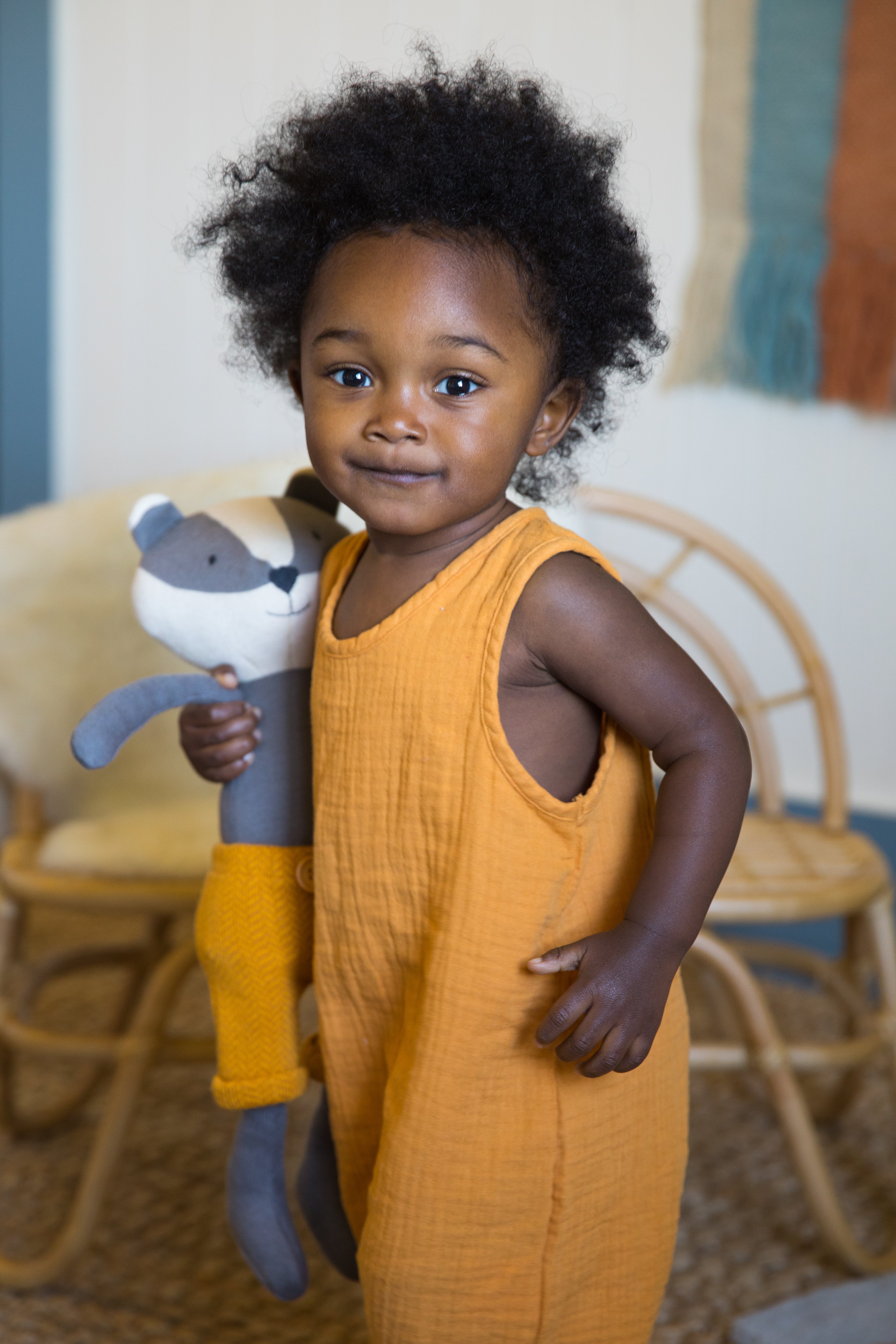 Baby wearing mustard organic muslin sleeveless coverall by Apple Park with soft knit toy